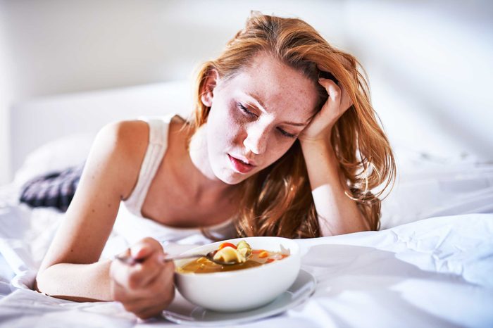 Woman lying on a bed eating a bowl of chicken soup and holding her forehead with her left hand.