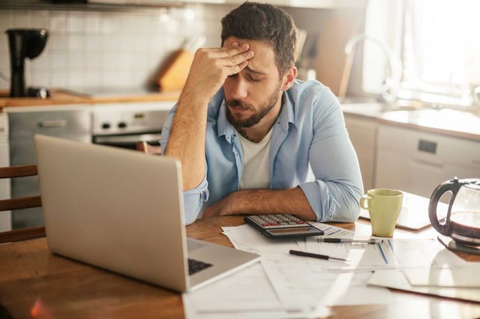 Man sitting at a kitchen table with his laptop and his hand on his forehead.