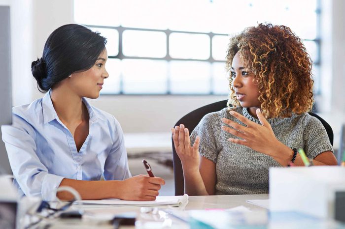 Two women office workers speaking to each other at a table.