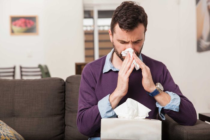 Man sitting on a couch blowing his nose with a tissue.