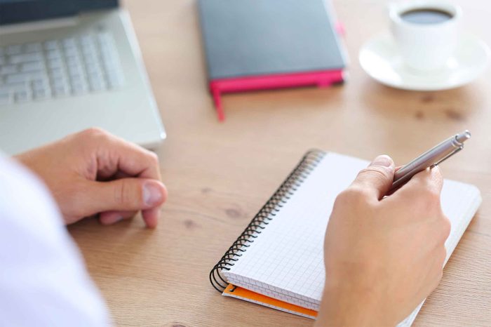 Person at a desk writing in a notebook with a pen.