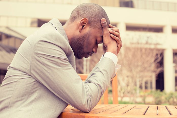 Black man sitting outdoors at a table, holding his head with his eyes closed