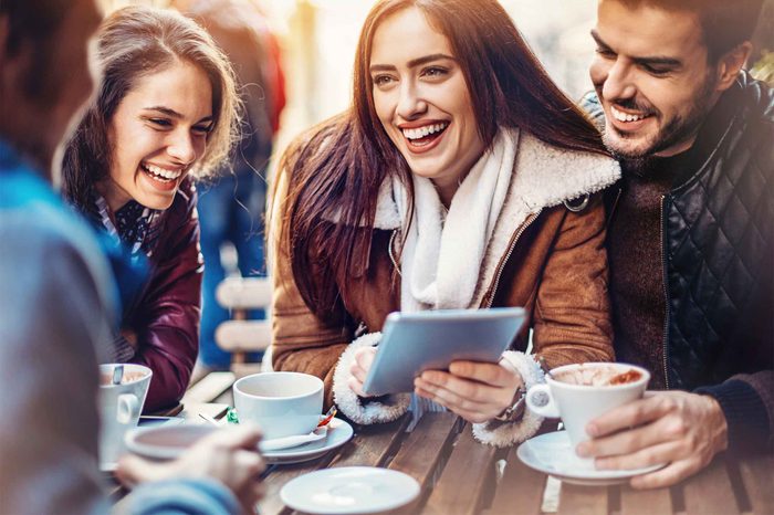 Woman wearing a shearling jacket holding a tablet while surrounded by friends at a table.