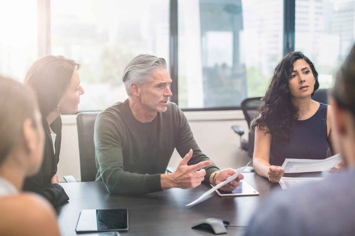 Man with gray hair sitting at a boardroom table holding a tablet surrounded by his co-workers.