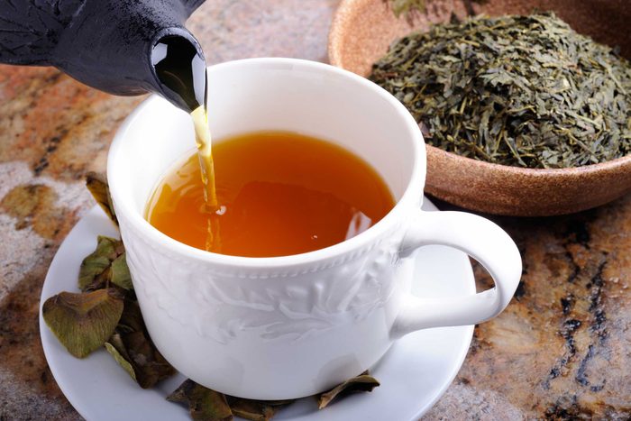 tea pot pouring into white mug and saucer next to bowl of tea leaves
