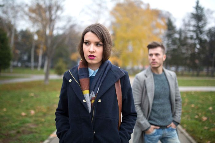 Woman wearing a coat and colorful scarf walking through a park as a man in a coat walks behind her.