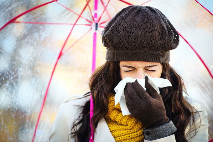 woman bundled up under an umbrella sneezing into a tissue