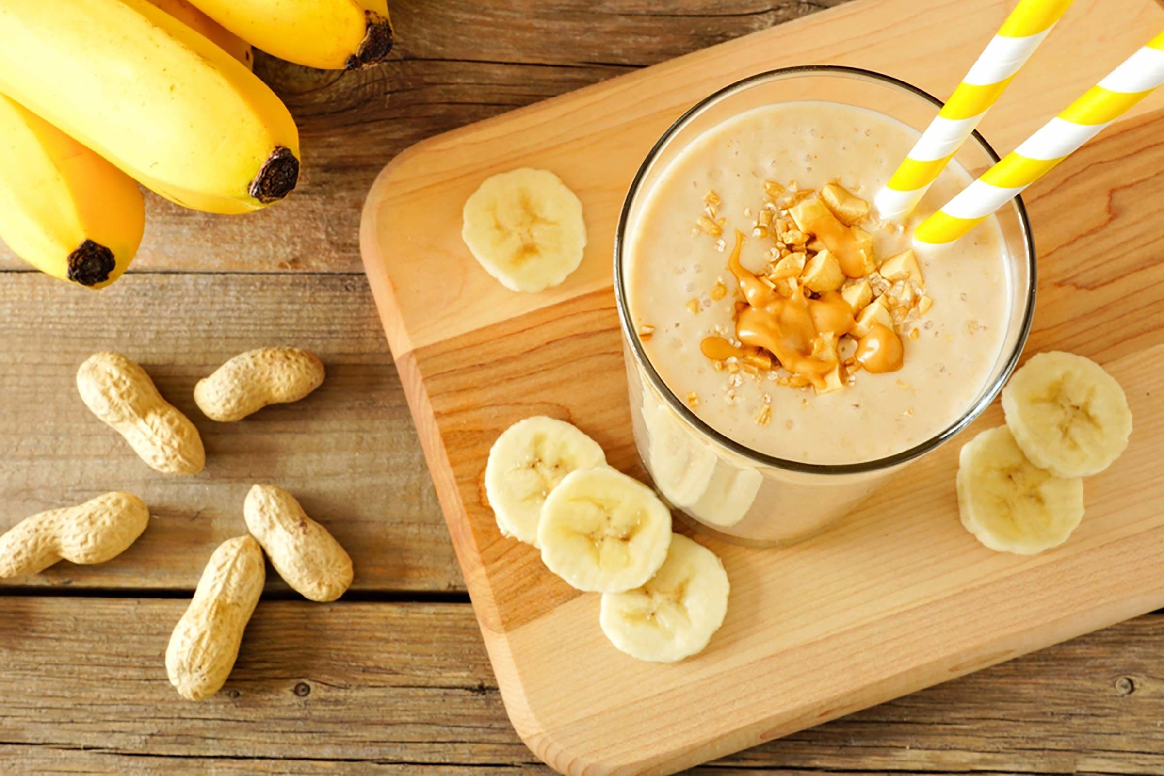 smoothie in a tall glass surrounded by banana slices, bananas, and peanuts in the shell