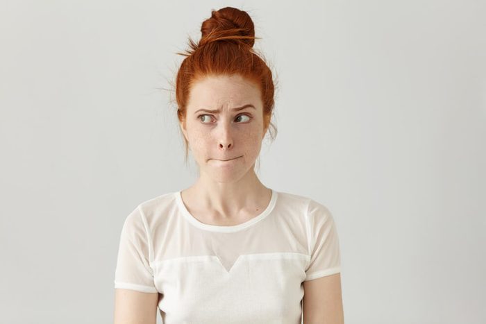 Indoor shot of cute redhead girl looking away, having doubtful and indecisive face expression, pursuing her lips as if forbidden to say anything. Confused young female posing isolated at white wall