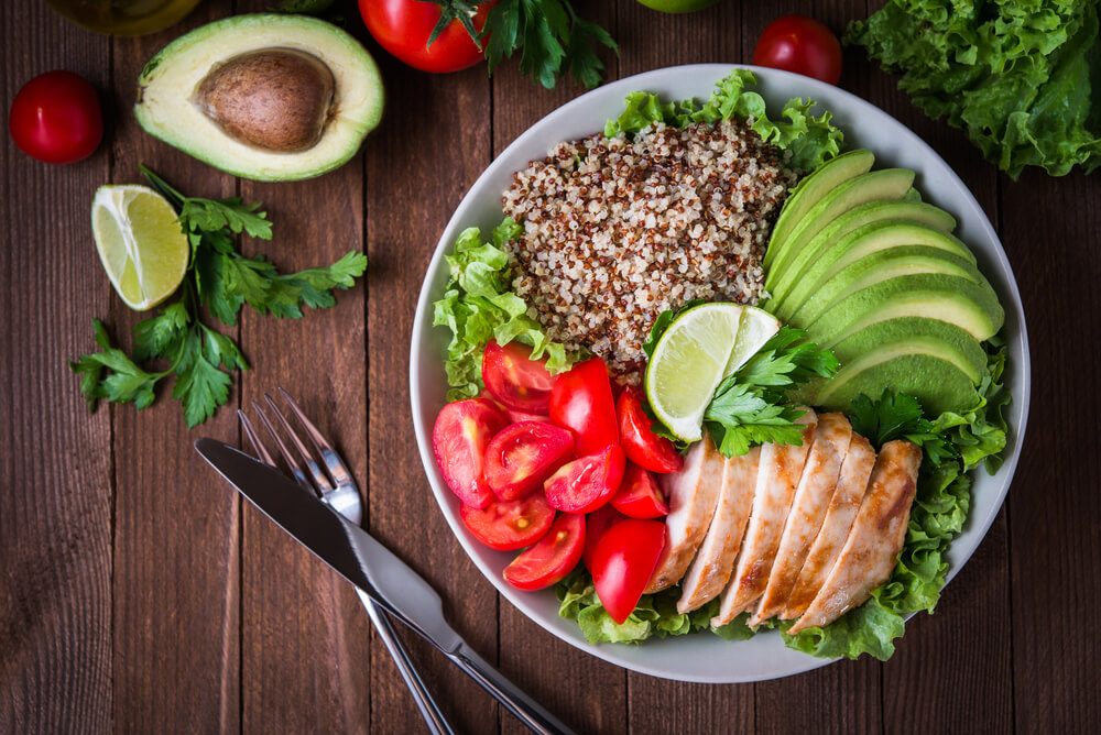 Healthy salad bowl with quinoa, tomatoes, chicken, avocado, lime and mixed greens (lettuce, parsley) on wooden background top view. Food and health.