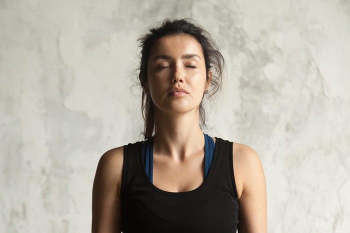 Portrait of young attractive yogi woman with her eyes closed in meditating pose, relaxation exercise, working out, wearing sportswear, black top, indoor close up image, wall background