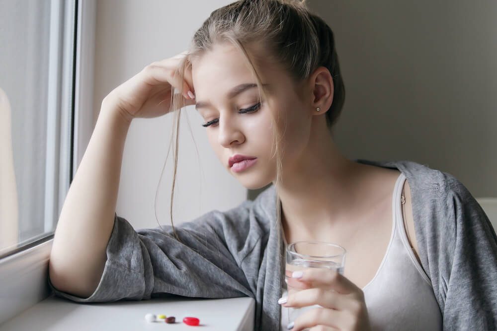 Displeased tired young beautiful girl or woman look on a pill or tablet with a glass of water near the window in white shirt and grey robe