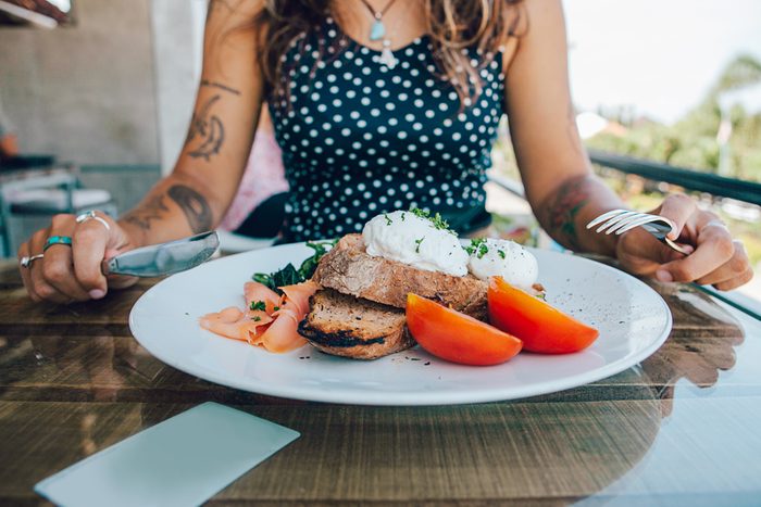 Poached eggs benedict served on toast with smoked salmon, avocado, grilled tomato and spinach, young woman eat breakfast with knife and fork
