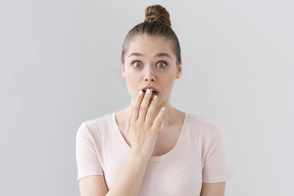 Indoor picture of surprised European female in casual outfit without make up isolated on gray background, looking astonished and deeply shocked, slightly covering round open mouth with hand.