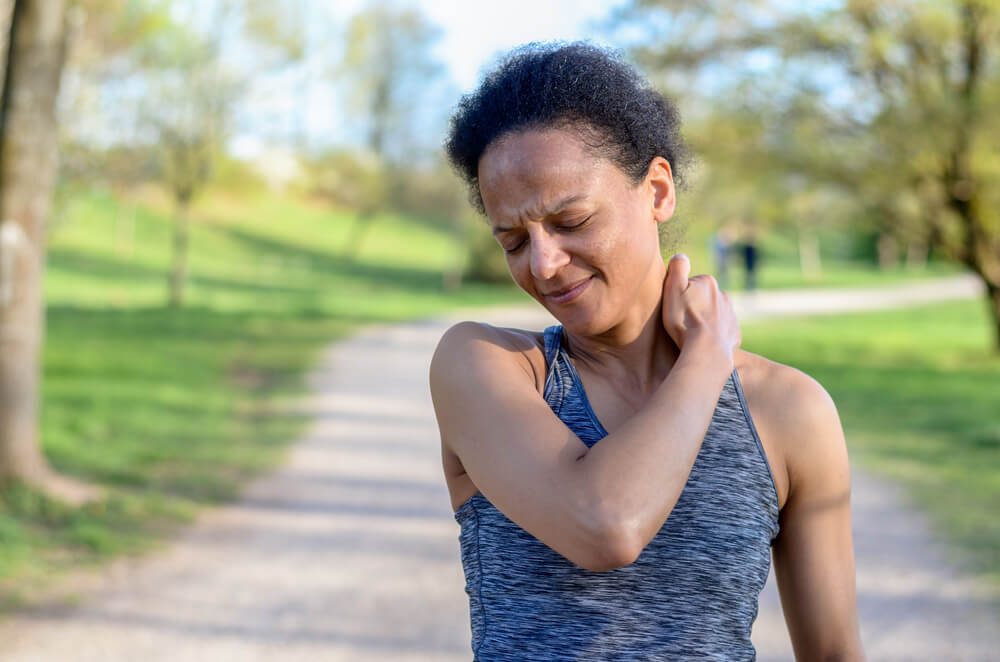 Young woman out jogging suffers a muscle injury standing holding her neck and lower back while grimacing in pain on a rural road, close up upper body view