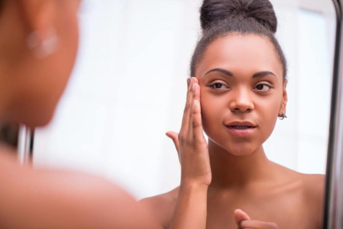 Beautiful dark skinned girl in white towel bringing face cream looking at mirror isolated on white background
