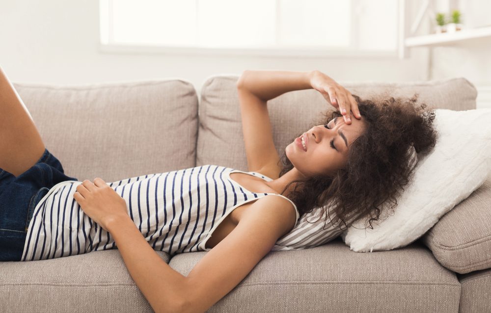 Sad girl with headache. Young african-american woman feeling pain, lying on sofa at home, copy space