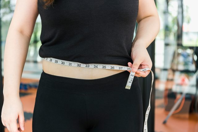 Woman measuring her waistline fat tummy on fitness center background,obese women,Body part of a fat woman with measuring tape