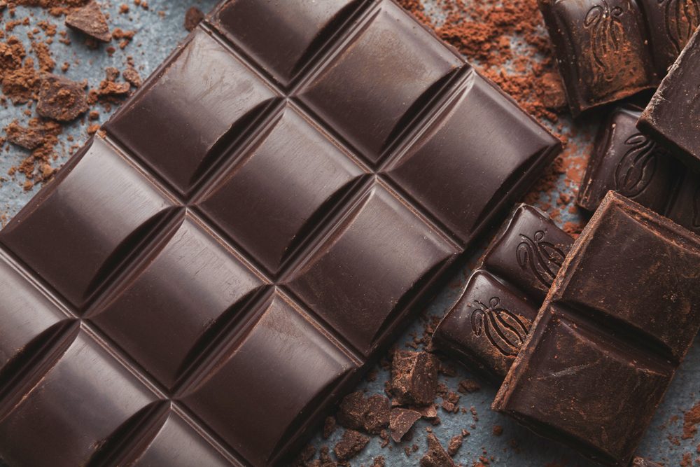 Dark chocolate bar broken in pieces on dark background and cocoa powder closeup. Sweet wallpaper, confectionery shop advertising and cooking ingredients concept