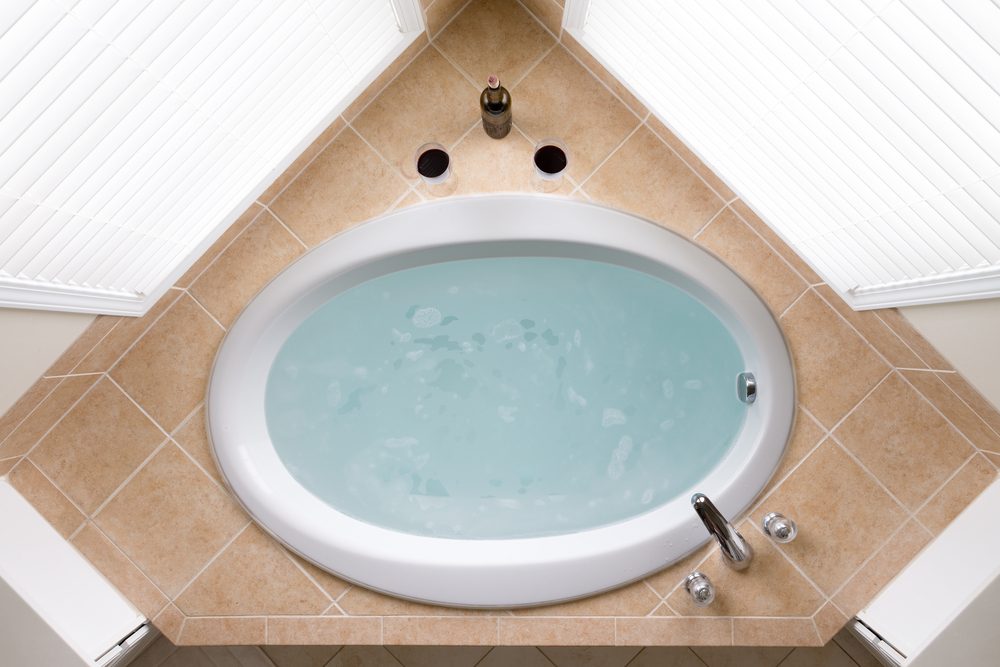 Stylish corner oval bathtub filled with fresh clean water in a brown tile surround flanked by two windows, overhead view