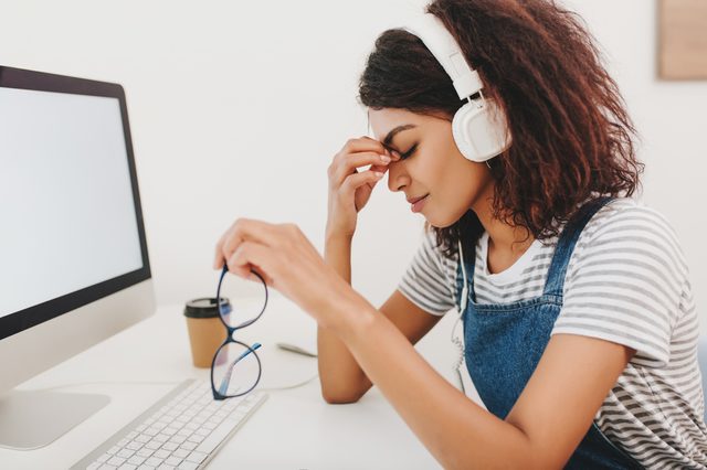 Graceful black woman in headphones with headache sitting beside computer and holding glasses in hand. Indoor portrait of tired female secretary posing with eyes closed at workplace.