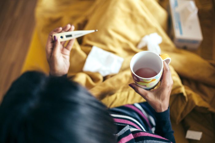 Sick woman drinking tea holding thermometer on couch