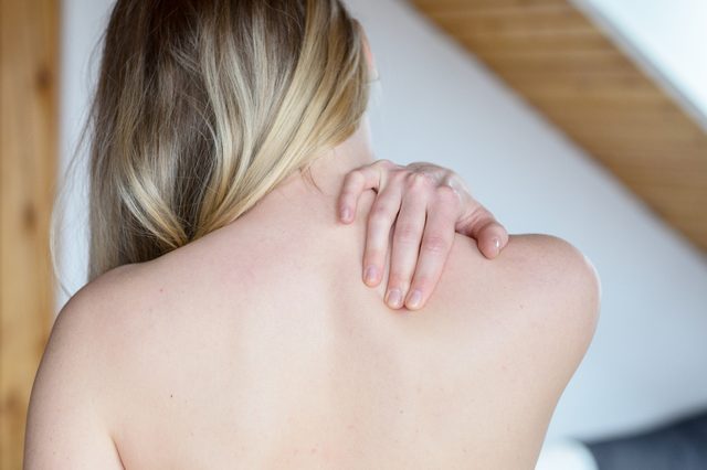 Rear upper body view of young woman rubbing painful shoulder with hand