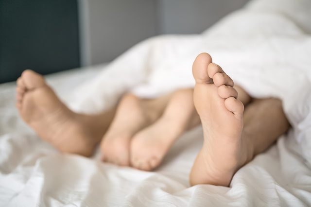 Closeup photo of the barefoot legs of lovers under the white blanket on the bed. Indoors. Closeup. Horizontal.