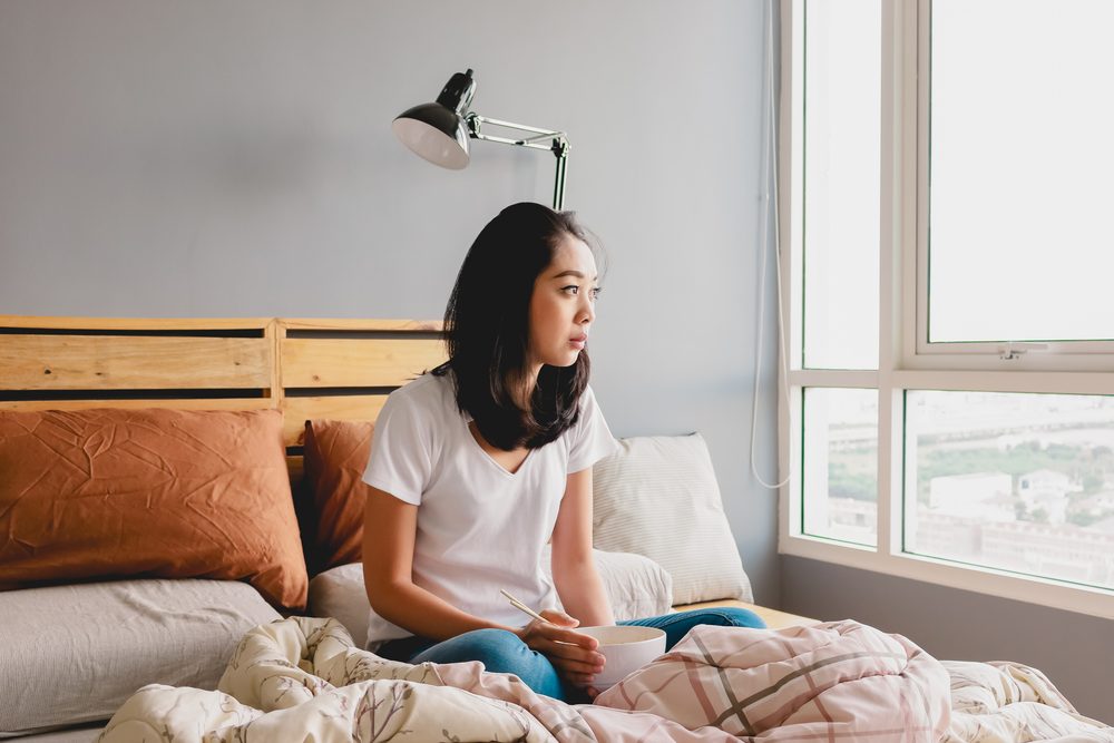 woman looking sad sitting and eating in bed, looking out the window