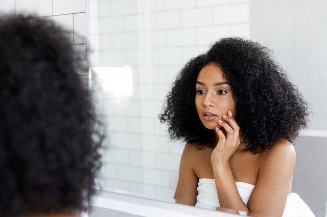 Attractive young woman touching her face, inspecting skin in front of the bathroom mirror