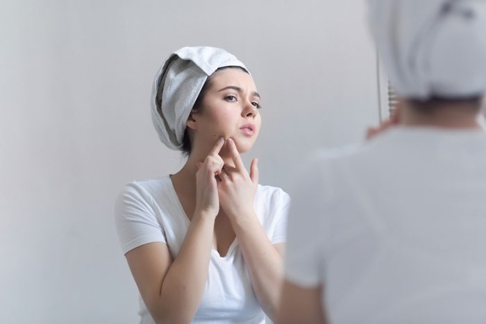 Young woman with acne in bathroom. Portrait of teenager girl looking at the mirror on her skin. Beauty, skin care lifestyle concept.