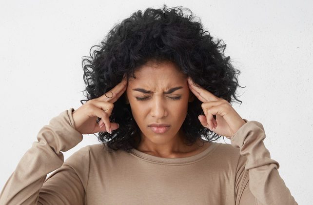 Dark-skinned young woman in beige long sleeved t-shirt frowning, having painful expressions, suffering from bad severe headache, keeping eyes closed, holding fingers on temples, trying to ease tension