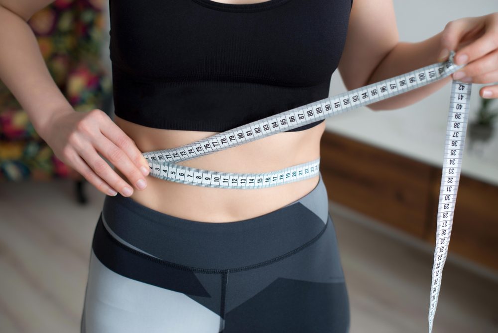A young woman in a sportswear checking success of weight loss program with measuring tape in front of mirror at home.
