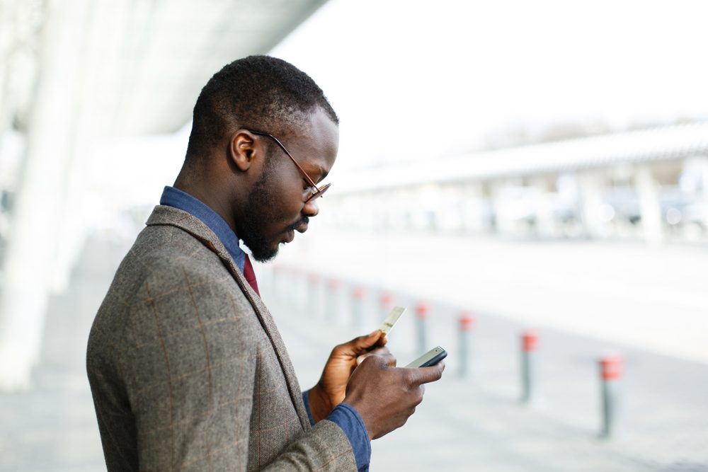 Stylish African American black businessman types information from his credit card to the smartphone standing before a modern glass building outside