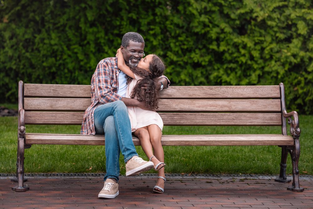 adorable african american girl hugging and kissing her grandfather while sitting on bench in park
