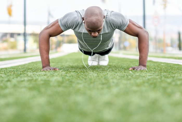 Man exercising push ups listening to music with headphones. Male model cross-training in urban background. African guy in his twenties doing workout outdoors in the street.