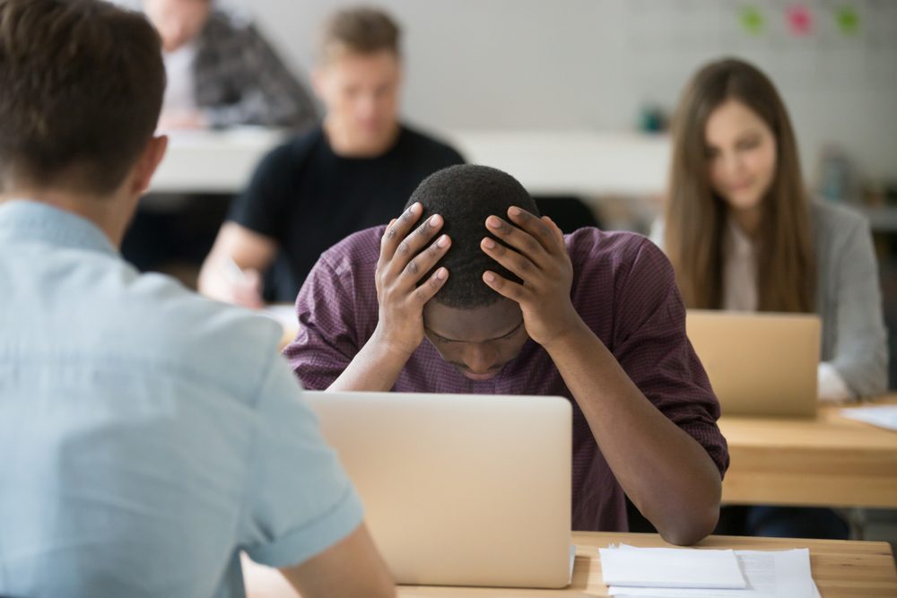 Frustrated african office worker having work problem holding head in hands sitting at desk in co-working with colleagues, depressed fired employee feeling hopeless distraught about corporate trouble