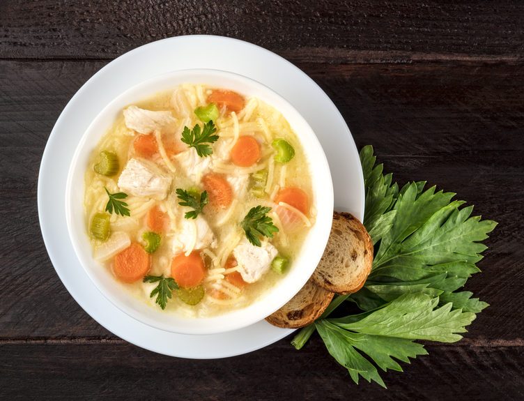 An overhead photo of a plate of chicken, vegetables, and noodles soup, shot from above on a dark rustic texture with slices of bread, a celery branch, and a place for text