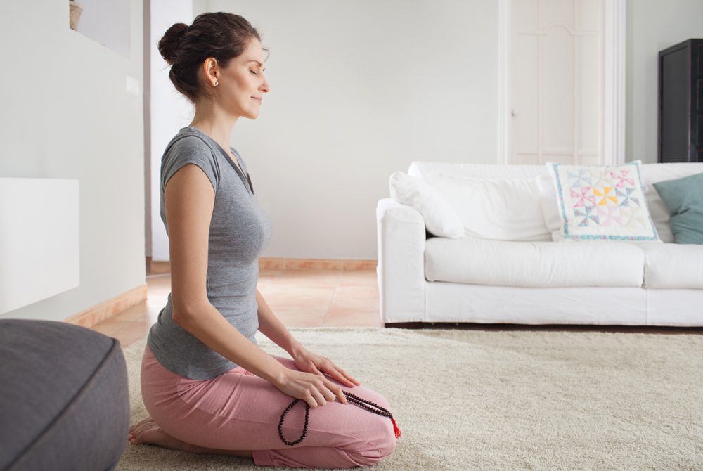 Side view of middle aged woman kneeling on carpet doing yoga with beads collar, home living room interior. Healthy female meditating, mind wellness mindful sport, indoors. Recreation lifestyle.