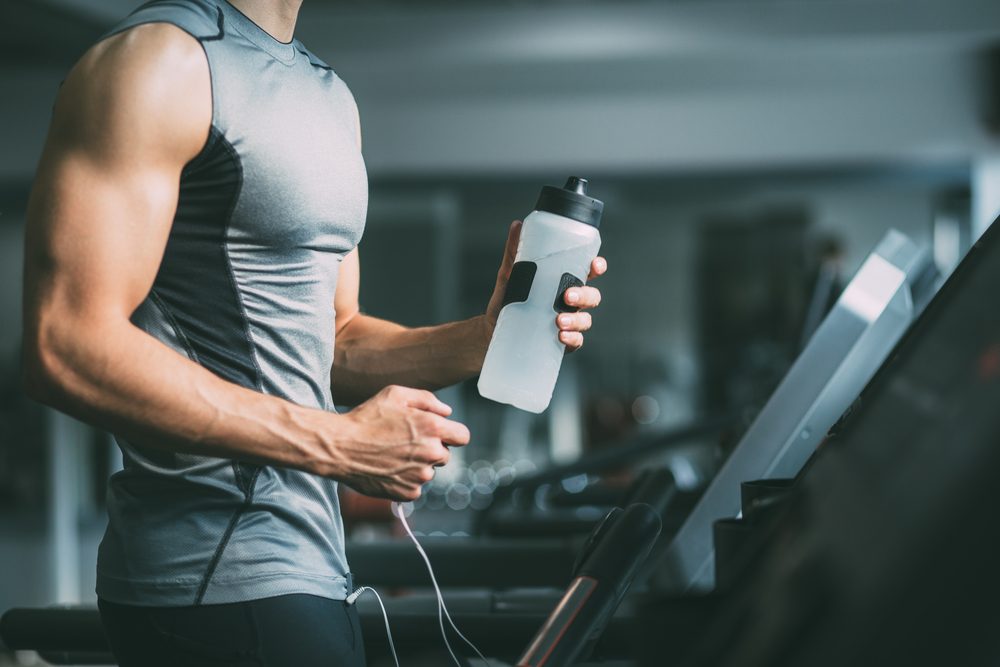 Unrecognizable young man in sportswear running on treadmill at gym and holding bottle of water