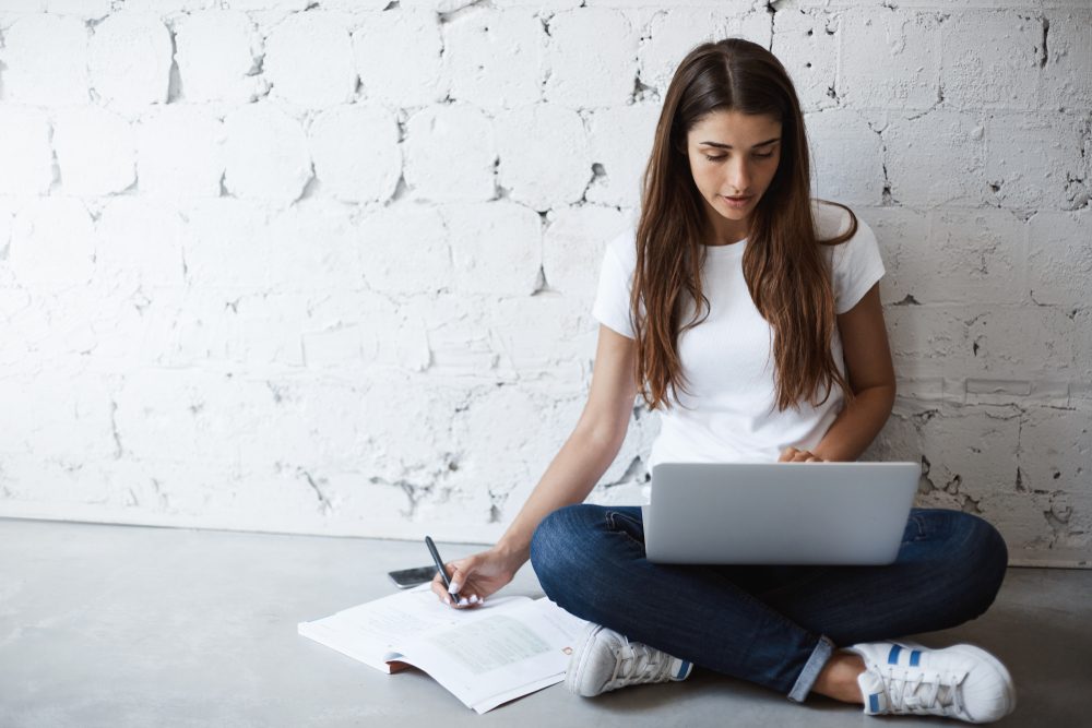 Indoor portrait of smart busy woman, making notes while leaning on wall and sitting on floor with laptop on crossed feet. Girl has habit to work or study everywhere but not at table. Study and work