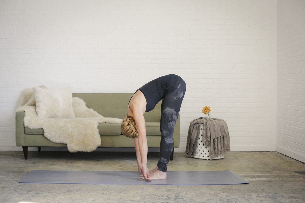 A blonde woman in a black leotard and leggings, standing on a yoga mat in a room, doing yoga, bending down, touching her toes with her hands