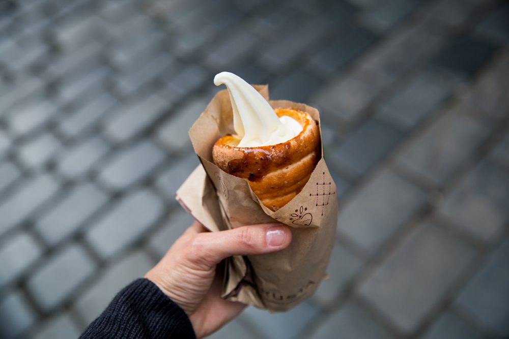 Tourist holds in hand Trdlo or Trdelnik with ice-cream on the background of city tourist streets of Czech (Prague). Fresh Appetizing Trdlo or Trdelnik - Traditional National Czech Sweet Pastry Dough.