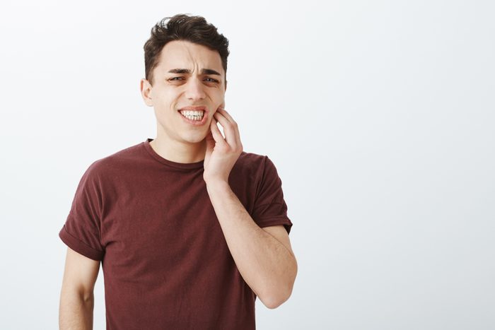 Portrait of displeased suffering young male model in red t-shirt, frowning and clenching fists, touching jaw while feeling toothache, having decay and dental problems, standing over gray background