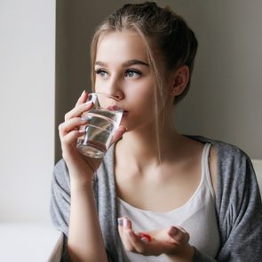 Young beautiful girl or woman take  a pill or tablet with a glass of water near the window in white shirt and grey robe