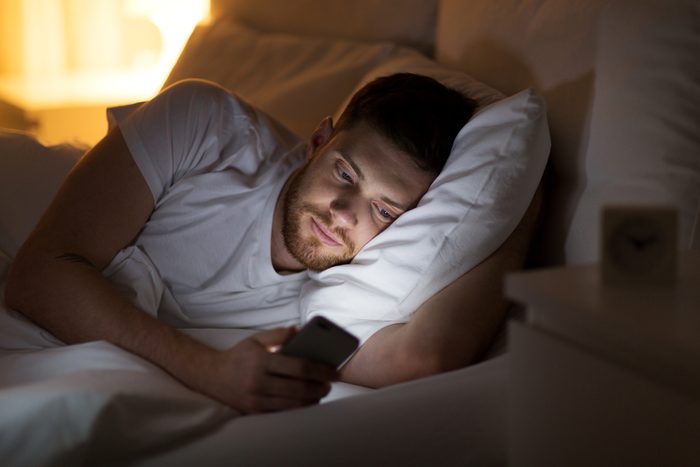 technology, internet, communication and people concept - happy smiling young man texting on smartphone in bed at home at night