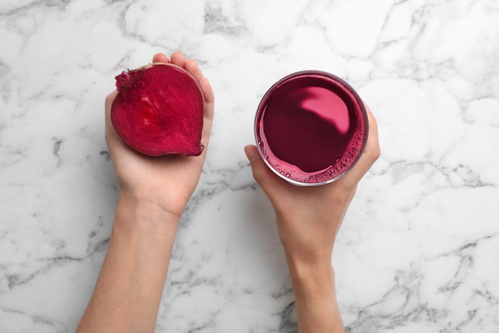 Woman holding glass with fresh beet juice and half of vegetable on marble table, top view