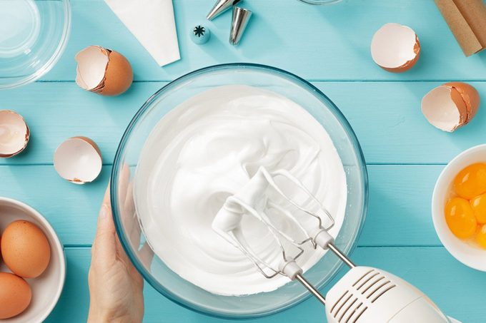 Whipped egg whites cream to perfect peaks in glass bowl with mixer and hand on blue wooden table.