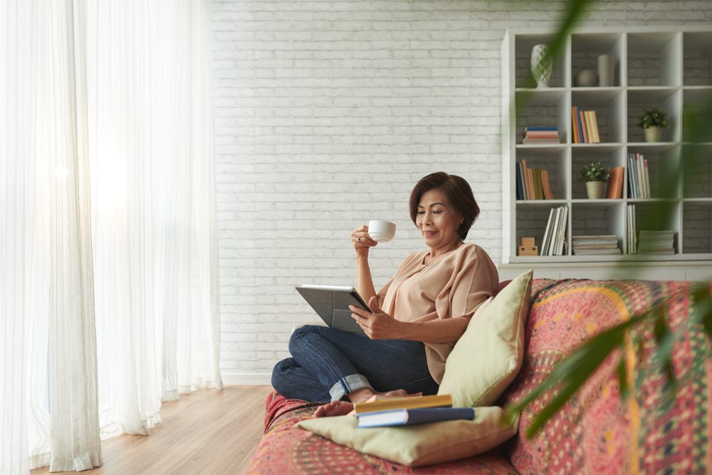 Vietnamese senior woman drinking tea and reading book on tablet computer