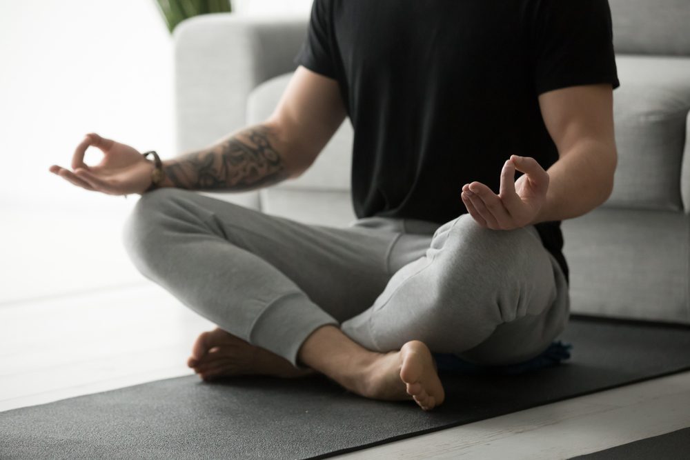 Sporty mindful man with tattoo meditating alone at home, peaceful calm hipster fit guy practicing yoga in lotus pose indoors holding hands in mudra, freedom and calmness concept, close up view